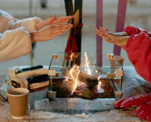 Two people warming hands over a glass-enclosed outdoor fire on a snowy day.